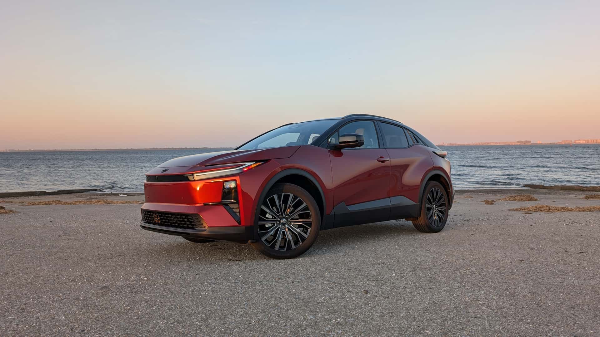 A red 2026 Toyota C-HR BEV parked on a gravel shoreline at sunset.